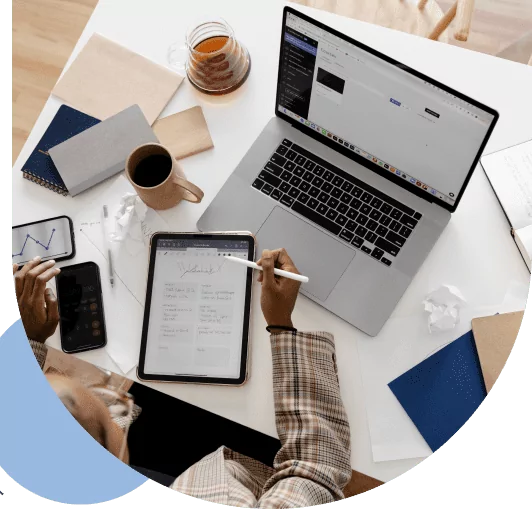 Desk from a birds-eye view, showing a tablet, laptop, tea, coffee, mobile phone, folders, and a notebook. A person's arm and lefthand is pictured using the tablet and phone.